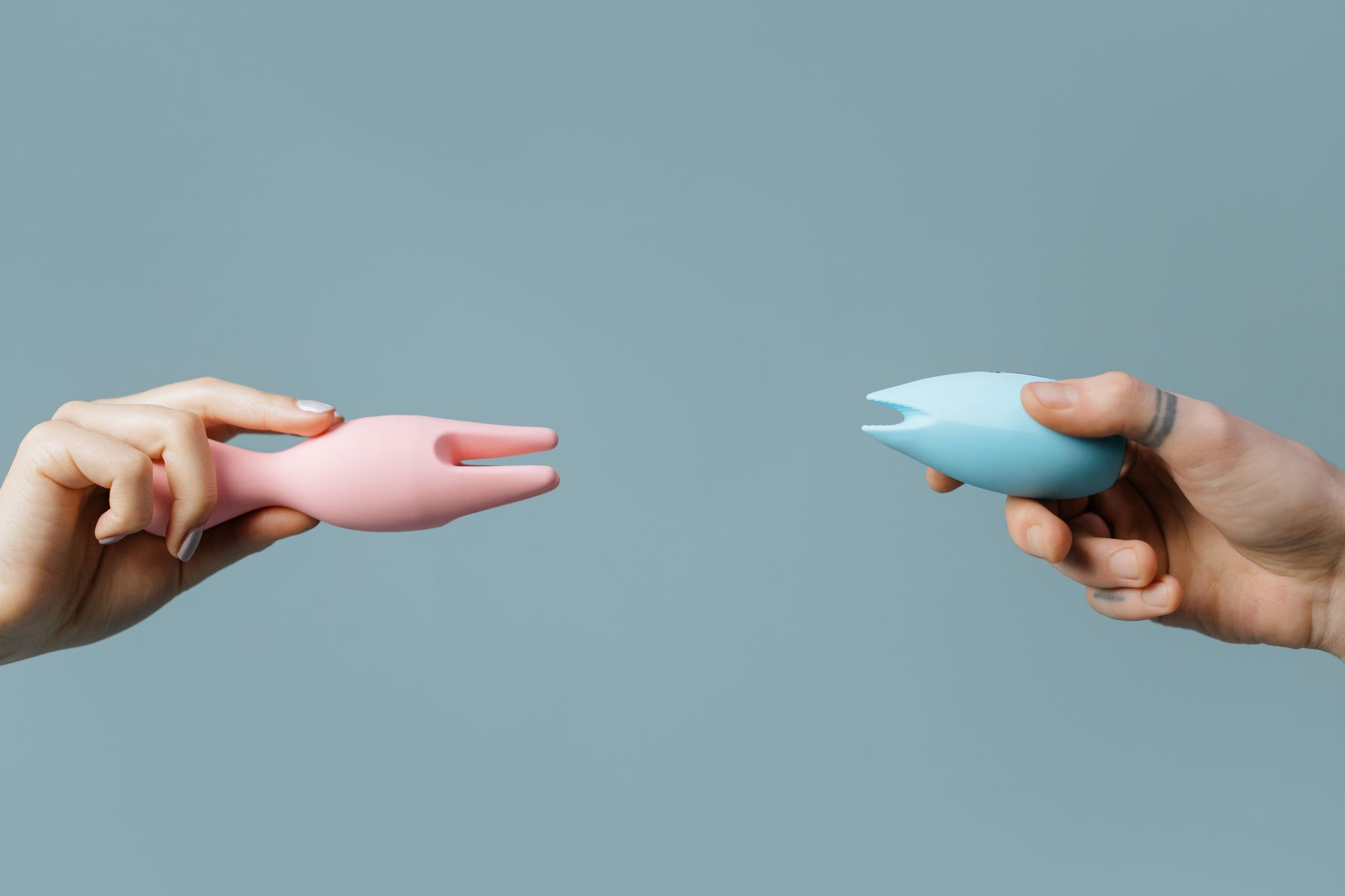 Two hands holding pastel-colored silicone vibrators against a soft background, representing shared pleasure and intimacy.